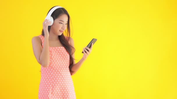 黄色に隔離されたスマートフォンやヘッドフォンで音楽を聴く美しいアジアの女性の映像 — ストック動画
