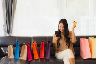 Güzel Asyalı bir kadının portresi. İnternetten alışveriş yapıyor. Kredi kartı ve akıllı telefonuyla oturma odasındaki alışveriş çantasının etrafında.