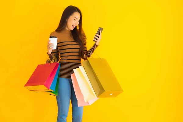 美丽的年轻亚洲女人使用智能手机或带有咖啡杯和黄色独立背景购物袋的手机 — 图库照片