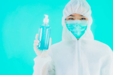 Güzel bir portre genç Asyalı doktor. Özel süit ve maske takıyor. Alkol jeli ve mavi izole bir arka plan.