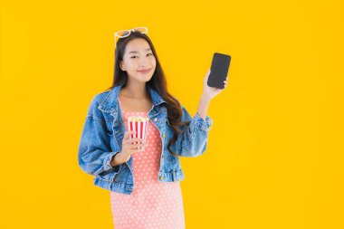 Güzel bir Asyalı kadın portresi telefon patlamış mısırı ve sarı izole edilmiş bir arka planda film izlemekten memnun.