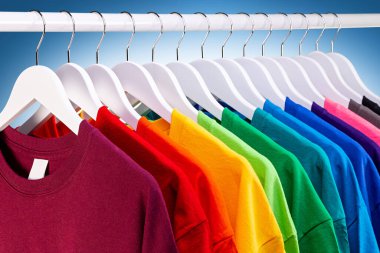 Renkli gökkuşağı renklerinde bir sürü yeni kumaş pamuk t-shirt 'ler gardıropta asılı. Mavi beyaz arka planda çeşitli renkli gömlekler. diy printing moda konsepti.
