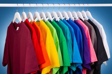 Renkli gökkuşağı renklerinde bir sürü yeni kumaş pamuk t-shirt 'ler gardıropta asılı. Mavi beyaz arka planda çeşitli renkli gömlekler. diy printing moda konsepti.