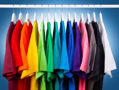 Renkli gökkuşağı renklerinde bir sürü yeni kumaş pamuk t-shirt 'ler gardıropta asılı. Mavi beyaz arka planda çeşitli renkli gömlekler. diy printing moda konsepti.