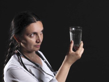 Kadın bir doktor elinde bir bardak su ve ilaç tutuyor ve duygusal olarak siyah bir arkaplanda poz veriyor.
