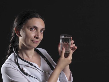 Kadın bir doktor elinde bir bardak su ve ilaç tutuyor ve duygusal olarak siyah bir arkaplanda poz veriyor.