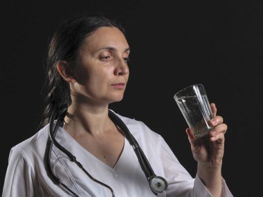 Kadın bir doktor elinde bir bardak su ve ilaç tutuyor ve duygusal olarak siyah bir arkaplanda poz veriyor.