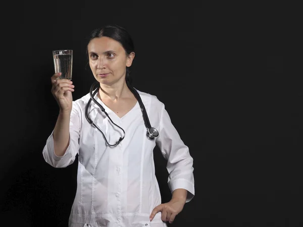 Uma médica feminina segura um copo de água e medicina e posa emocionalmente em um fundo preto — Fotografia de Stock
