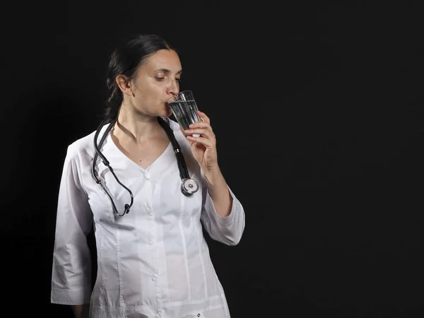Un medico donna tiene un bicchiere d'acqua e medicina e posa emotivamente su uno sfondo nero — Foto Stock