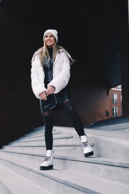 Uzun saçlı sarışın, beyaz yün şapkalı, altında kıvrımlı siyah deri bir elbise olan beyaz ceketli, siyah çoraplı ve beyaz çizmeli, zarif bir şekilde merdivenlerden aşağı iniyor.