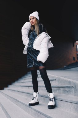 Uzun saçlı sarışın, beyaz yün şapkalı, altında kıvrımlı siyah deri bir elbise olan beyaz ceketli, siyah çoraplı ve beyaz çizmeli, zarif bir şekilde merdivenlerden aşağı iniyor.