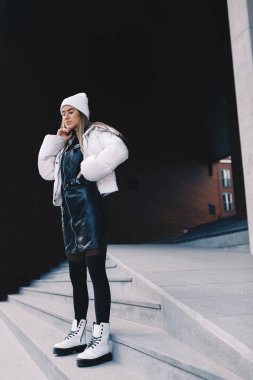 Uzun saçlı sarışın, beyaz yün şapkalı, altında kıvrımlı siyah deri bir elbise olan beyaz ceketli, siyah çoraplı ve beyaz çizmeli, zarif bir şekilde merdivenlerden aşağı iniyor.