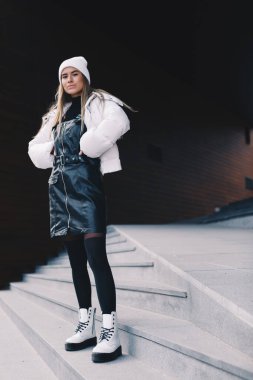 Uzun saçlı sarışın, beyaz yün şapkalı, altında kıvrımlı siyah deri bir elbise olan beyaz ceketli, siyah çoraplı ve beyaz çizmeli, zarif bir şekilde merdivenlerden aşağı iniyor.