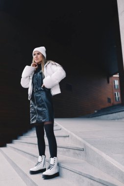 Uzun saçlı sarışın, beyaz yün şapkalı, altında kıvrımlı siyah deri bir elbise olan beyaz ceketli, siyah çoraplı ve beyaz çizmeli, zarif bir şekilde merdivenlerden aşağı iniyor.