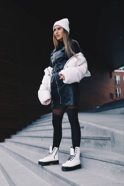 Uzun saçlı sarışın, beyaz yün şapkalı, altında kıvrımlı siyah deri bir elbise olan beyaz ceketli, siyah çoraplı ve beyaz çizmeli, zarif bir şekilde merdivenlerden aşağı iniyor.