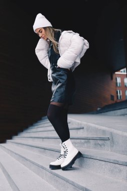 Uzun saçlı sarışın, beyaz yün şapkalı, altında kıvrımlı siyah deri bir elbise olan beyaz ceketli, siyah çoraplı ve beyaz çizmeli, zarif bir şekilde merdivenlerden aşağı iniyor.