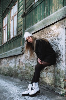 Sarışın bir kadın, taş bir duvara yaslanmış, beyaz yün bir şapka, siyah çoraplar giyiyor, beyaz çizmeler, önümdeki günü düşünüyor.