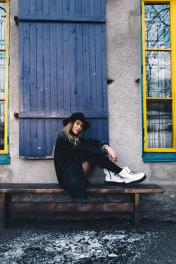 Uzun saçlı, sarı saçlı, siyah fötr şapka takan siyah yakalı bir kadın ve mavi pencerenin arkasındaki ahşap bankta oturan beyaz çizmeler.