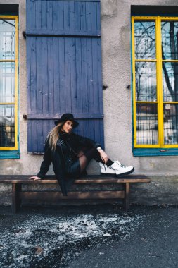 Uzun saçlı, sarı saçlı, siyah fötr şapka takan siyah yakalı bir kadın ve mavi pencerenin arkasındaki ahşap bankta oturan beyaz çizmeler.