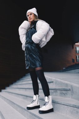 Uzun saçlı sarışın, beyaz yün şapkalı, altında kıvrımlı siyah deri bir elbise olan beyaz ceketli, siyah çoraplı ve beyaz çizmeli, zarif bir şekilde merdivenlerden aşağı iniyor.