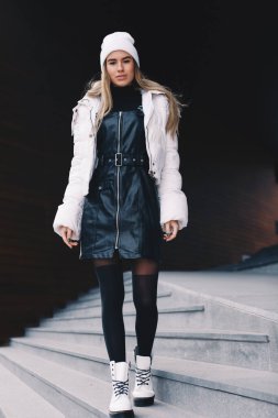 Uzun saçlı sarışın, beyaz yün şapkalı, altında kıvrımlı siyah deri bir elbise olan beyaz ceketli, siyah çoraplı ve beyaz çizmeli, zarif bir şekilde merdivenlerden aşağı iniyor.