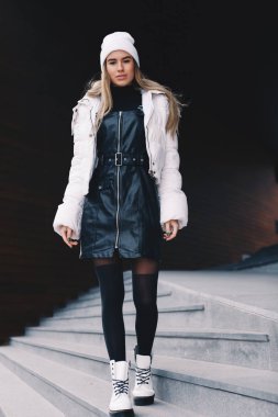 Uzun saçlı sarışın, beyaz yün şapkalı, altında kıvrımlı siyah deri bir elbise olan beyaz ceketli, siyah çoraplı ve beyaz çizmeli, zarif bir şekilde merdivenlerden aşağı iniyor.