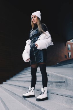 Uzun saçlı sarışın, beyaz yün şapkalı, altında kıvrımlı siyah deri bir elbise olan beyaz ceketli, siyah çoraplı ve beyaz çizmeli, zarif bir şekilde merdivenlerden aşağı iniyor.