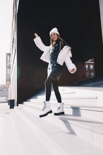 Long haired blonde, wearing a white woollen cap, a white jacket with a curvaceous black leather dress underneath, black stockings and white boots, walking gracefully down a flight of stairs