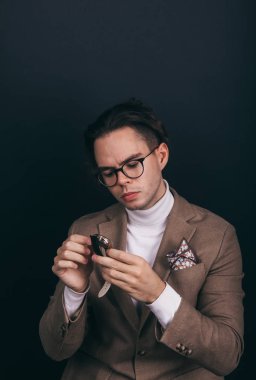 A dapper young man obviously you wearing a white turtleneck sweater and a beige jacket with handkerchief in his top breast pocket, looking pensively at his wrist watch, contemplating time