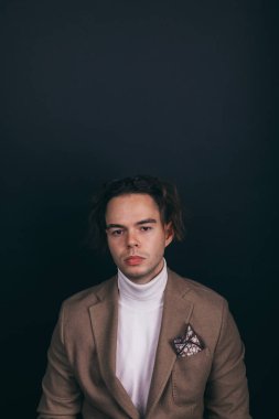 A dapper young man obviously you wearing a white turtleneck sweater and a beige jacket with handkerchief in his top breast pocket, looking pensively at his wrist watch, contemplating time
