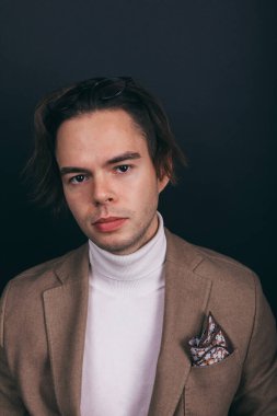 A dapper young man obviously you wearing a white turtleneck sweater and a beige jacket with handkerchief in his top breast pocket, looking pensively at his wrist watch, contemplating time