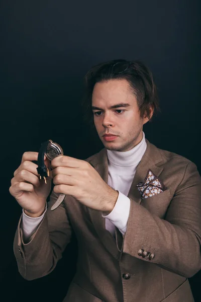 A dapper young man obviously you wearing a white turtleneck sweater and a beige jacket with handkerchief in his top breast pocket, looking pensively at his wrist watch, contemplating time — ストック写真