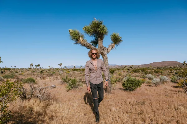 Joshua tree a curly haired blonde man is walking in the fierce heat and deep blue skies of the south western desert of North America, wearing dark sunglasses, a beige linen shirt, and surprisingly bla — Stock Photo, Image
