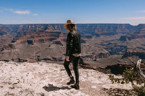 A Curly haired blonde man, wearing a black leather jacket ,black jeans, black shoes, beige linen shirt and matching cowboy hat, background vistas of the Grand Canyon — Stock Photo, Image