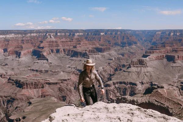 Ein blonder Mann mit lockigem Haar, bekleidet mit beigem T-Shirt, schwarzer Jeans, schwarzen Schuhen, beigem Leinenhemd und passendem Cowboyhut, Blick auf den Grand Canyon — Stockfoto