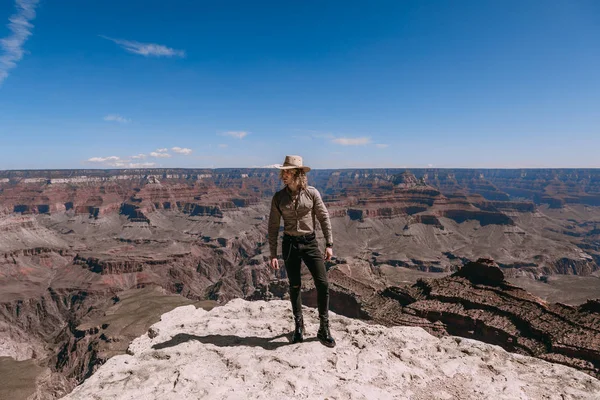 Ein blonder Mann mit lockigem Haar, bekleidet mit beigem T-Shirt, schwarzer Jeans, schwarzen Schuhen, beigem Leinenhemd und passendem Cowboyhut, Blick auf den Grand Canyon — Stockfoto