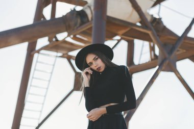 Siyah şapkalı, siyah fötr şapka takan, uzun saçlı, sarışın bir kadın. Siyah güneş gözlüklü, diz boyu siyah bir elbise. Sıkıntılı bir endüstriyel silonun önünde utanmadan poz veriyor.