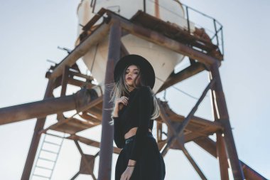 Siyah şapkalı, siyah fötr şapka takan, uzun saçlı, sarışın bir kadın. Siyah güneş gözlüklü, diz boyu siyah bir elbise. Sıkıntılı bir endüstriyel silonun önünde utanmadan poz veriyor.