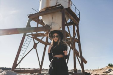 Siyah şapkalı, siyah fötr şapka takan, uzun saçlı, sarışın bir kadın. Siyah güneş gözlüklü, diz boyu siyah bir elbise. Sıkıntılı bir endüstriyel silonun önünde utanmadan poz veriyor.