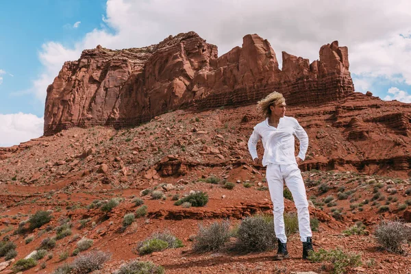 A curly haired blonde man posing around the famous Buttes of Monument Valley from Arizona, USA, wearing white linen shirt and white pants, black shoes, majestic beauty.