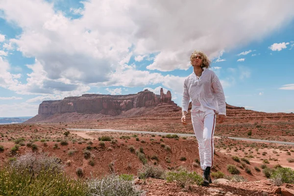 A curly haired blonde man posing around the famous Buttes of Monument Valley from Arizona, USA, wearing white linen shirt and white pants, black shoes, majestic beauty.