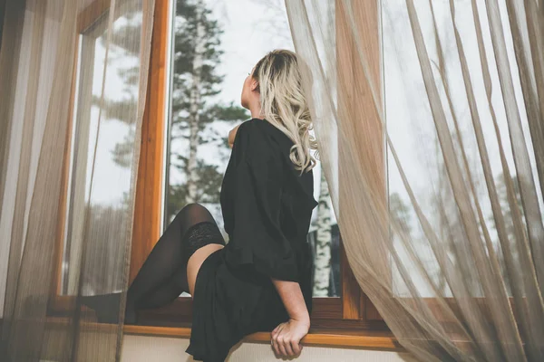 Mujer mirando a través de la vieja ventana sentada en el alféizar de la ventana. Vestido con medias — Foto de Stock