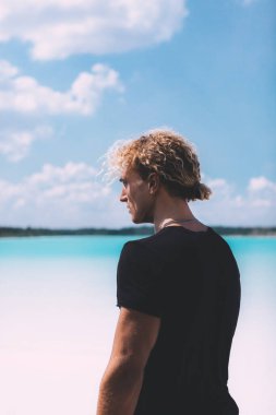 Açık kumsalda poz veren kıvırcık saçlı bir adam. Siyahlar içinde. Harika bir arka plan, temiz su ve gökyüzü. Moda, tropikal ve çekici bir adam. Yaz zamanı..