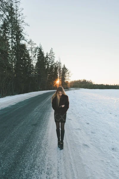 Young and alone girl dressed fur coat and dress standing around outdoor road. Sunset on the background. Sunlight and faded colors. Snow