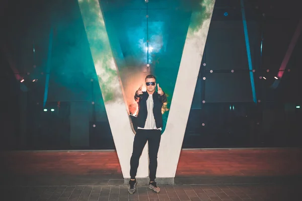 Man in a leather jacket standing at the street with smoke bomb. Colorful portrait of european teenager with red smoke bomb.