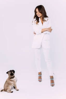 Beyaz elbiseli ve beyaz ayakkabılı, gülen ve gülümseyen kumral saçlı bir kadın, sakince oturup, tüm dikkatini toplayan sevimli köpeğine sevgiyle bakıyor. İzole edilmiş beyaz arkaplan