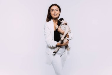 Beyaz elbiseli, gülen ve gülümseyen kumral saçlı bir kadın, sakince elleri üzerinde oturan ve tüm dikkatini toplayan sevimli köpeğine sevgiyle bakıyor. İzole edilmiş beyaz arkaplan