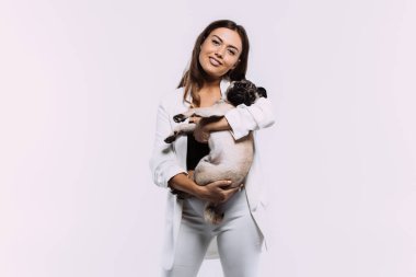 Beyaz elbiseli, gülen ve gülümseyen kumral saçlı bir kadın, sakince elleri üzerinde oturan ve tüm dikkatini toplayan sevimli köpeğine sevgiyle bakıyor. İzole edilmiş beyaz arkaplan
