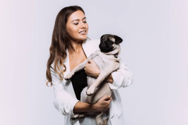 Beyaz elbiseli, gülen ve gülümseyen kumral saçlı bir kadın, sakince elleri üzerinde oturan ve tüm dikkatini toplayan sevimli köpeğine sevgiyle bakıyor. İzole edilmiş beyaz arkaplan