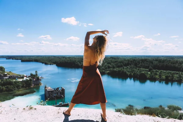 At a picturesque vantage point of Rummu quarry, a blond woman is standing in her bare feet, wearing a thigh high, auburn coloured dress, With her back turned away from the camera. Clear Water